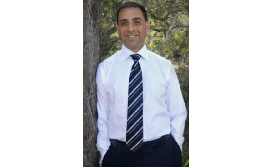 A man in a white shirt and striped tie stands outdoors, smiling confidently as if he just finished a refreshing session of hyperbaric chamber therapy, with lush greenery in the background.