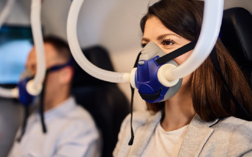 Two people experiencing Mild Hyperbaric Oxygen Therapy are seated in a transport vehicle, each wearing oxygen masks with attached tubes.